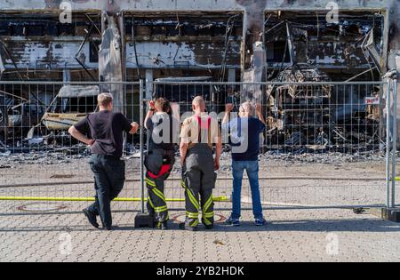 Stadtallendorf, Germania. 16 ottobre 2024. I vigili del fuoco stanno alla recinzione e guardano il capannone delle attrezzature bruciate. Il nuovo deposito di attrezzature del dipartimento dei vigili del fuoco volontario di Stadtallendorf è bruciato, completo di veicoli e attrezzature, causando milioni di danni. Crediti: Andreas Arnold/dpa/Alamy Live News Foto Stock