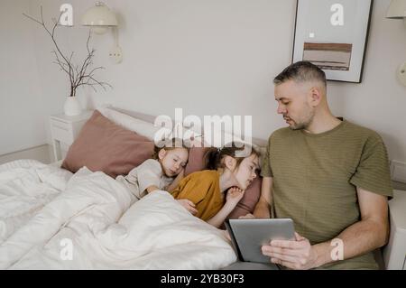 Un padre sta usando un tablet mentre le sue due figlie dormono pacificamente a letto. Sta guardando il tablet, magari guardando qualcosa di divertente o.. Foto Stock