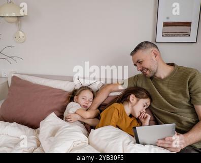 Un padre sta usando un tablet mentre le sue due figlie dormono a letto. Sta sorridendo e guardando il tablet, magari guardando qualcosa di divertente o.. Foto Stock