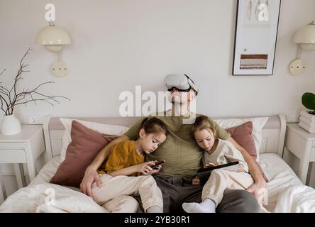 Un padre sta godendo della realtà virtuale mentre le sue due figlie si siedono accanto a lui, una su un tablet e l'altra su un telefono. Foto Stock