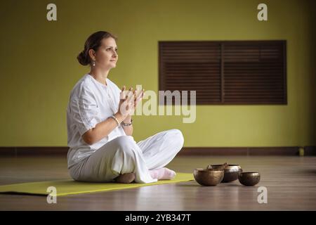 Donna che pratica yoga Kundalini, seduta in comoda posizione sul tappeto e che fa mudra di loto dalla solitudine, pratica in abiti bianchi con l'ingegno della camera Foto Stock