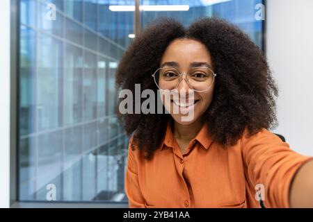 Donna sicura di sé con capelli ricci e occhiali sorridenti mentre scatta selfie in interni. Edificio luminoso che offre un ambiente fresco e professionale. Foto Stock