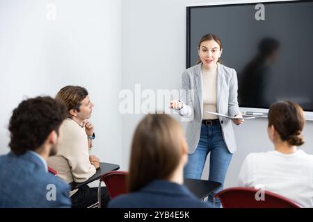 Giovane donna in piedi vicino a una lavagna interattiva e che comunica con gli studenti adulti durante i corsi di formazione avanzata in classe Foto Stock