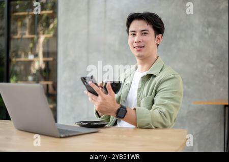 Un uomo asiatico bello e felice in abiti informali è seduto a un tavolo in una caffetteria, tenendo in mano uno smartphone e una tazza di caffè, con un computer portatile sul tavolo Foto Stock