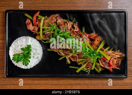 Maiale fritto con frecce all'aglio e verdure con riso su un piatto nero su un tavolo di legno Foto Stock