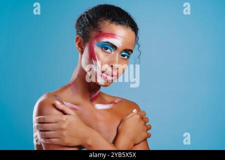 Studio, ritratto e donna con pittura per il viso, sorriso e colore del trucco, cosmetici e ombre per gli occhi. Sfondo blu, calma e art deco per la persona Foto Stock