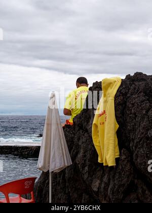 Un bagnino siede su un masso vulcanico, con una felpa gialla drappeggiata sulla pietra, che si affaccia sull'oceano presso le piscine naturali di Porto Moniz Foto Stock