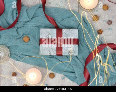 Regalo di Natale blu avvolto da un nastro Bordeaux. Coperta blu sullo sfondo. Decorata con candele, biglie, noci e luci sparse. Foto Stock