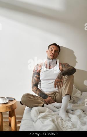 Un bel giovane uomo si siede su un letto, godendosi un momento tranquillo con un drink e un atteggiamento premuroso. Foto Stock