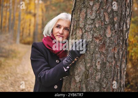 Una bella donna matura dai capelli argentati abbraccia l'albero nella foresta autunnale, godendo di foglie dorate che cadono, si rilassa e si riconnette con la natura, pacifica e Foto Stock