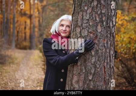 Una bella donna matura dai capelli argentati abbraccia l'albero nella foresta autunnale, godendo di foglie dorate che cadono, si rilassa e si riconnette con la natura, pacifica e Foto Stock