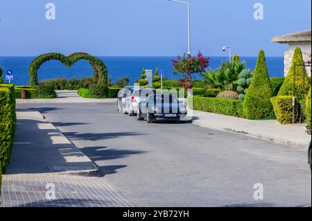 Un vivace sentiero costiero mostra eleganti siepi a forma di cuore, mentre le auto parcheggiano tranquillamente accanto al verde lussureggiante, incorniciate da un oceano blu brillante Foto Stock