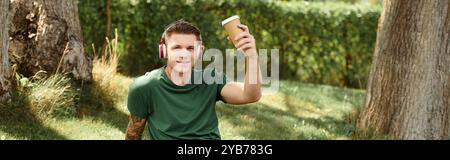 Un giovane uomo siede sull'erba, indossa le cuffie e sorride mentre tiene una tazza di caffè, abbracciando la vita e la gioia. Foto Stock