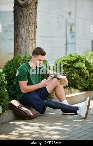 Un giovane con una gamba protesica siede a terra, sorseggiando un caffè e controllando il telefono in una posizione soleggiata. Foto Stock