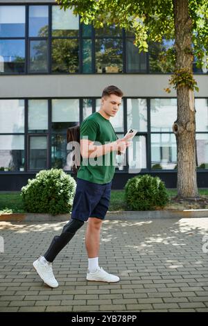 Un giovane e affascinante uomo con una gamba protesica cammina con sicurezza all'esterno, concentrandosi sul suo smartphone. Foto Stock