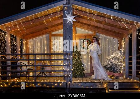 Donna Christmas Lights Cabin - Una donna con un abito bianco si erge sul portico di una cabina di legno decorata con luci natalizie. Foto Stock