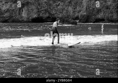 Una giovane surfista pratica il suo equilibrio su una piccola onda durante una lezione di surf alla spiaggia di Seixal, catturata in bianco e nero Foto Stock