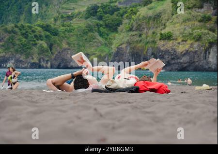 La coppia trascorre una giornata rilassante leggendo sulla sabbia nera della spiaggia di Seixal, con lussureggianti scogliere e acque turchesi a Madeira Foto Stock