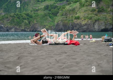 La coppia trascorre una giornata rilassante leggendo sulla sabbia nera della spiaggia di Seixal, con lussureggianti scogliere e acque turchesi a Madeira Foto Stock