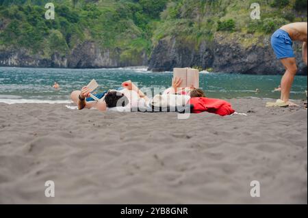 La coppia trascorre una giornata rilassante leggendo sulla sabbia nera della spiaggia di Seixal, con lussureggianti scogliere e acque turchesi a Madeira Foto Stock