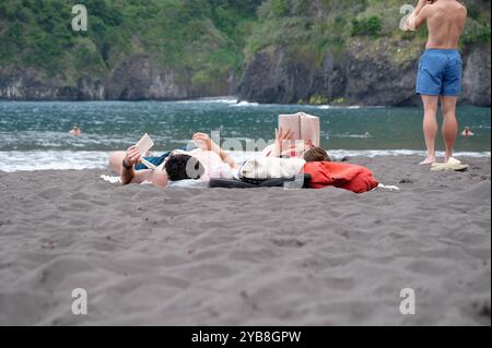 La coppia trascorre una giornata rilassante leggendo sulla sabbia nera della spiaggia di Seixal, con lussureggianti scogliere e acque turchesi a Madeira Foto Stock