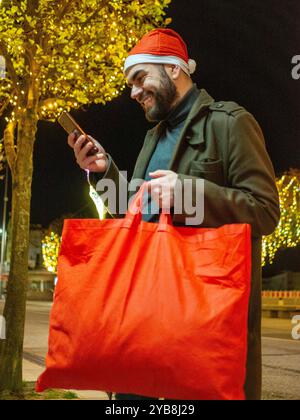 Uomo barbuto che usa il suo telefono mentre guarda la sua borsa della spesa durante il periodo natalizio Foto Stock