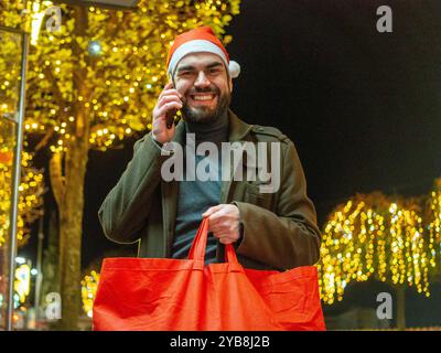 Uomo barbuto che usa il suo telefono mentre guarda la sua borsa della spesa durante il periodo natalizio Foto Stock