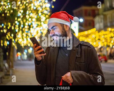 Uomo barbuto che usa il suo telefono mentre guarda la sua borsa della spesa durante il periodo natalizio Foto Stock