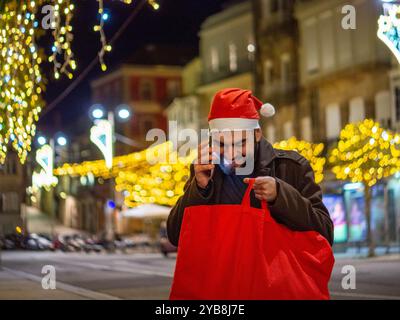 Uomo barbuto che usa il suo telefono mentre guarda la sua borsa della spesa durante il periodo natalizio Foto Stock