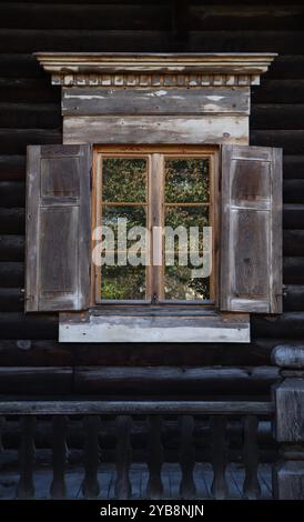 primo piano di una finestra in legno con persiane aperte sull'esterno di una cabina in legno scuro, struttura di legno invecchiato, artigianato tradizionale, riflesso di alberi verdi in vetro. finestra rustica, concetto di nostalgia Foto Stock