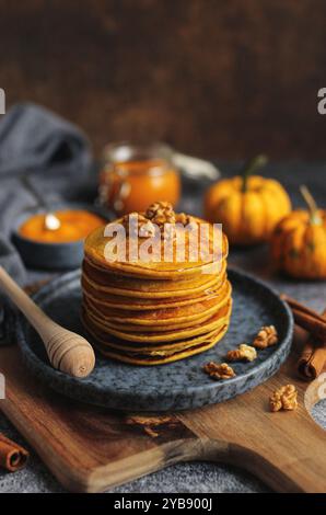 Pancake di zucca leggeri e soffici con miele e noci su un piatto blu. Tagliere in legno, zucca pura in un vaso di vetro, bastoncini di cannella e scura Foto Stock