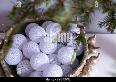Una collezione di sfere decorative bianche è disposta in una ciotola verde, parzialmente nascosta da rami sempreverdi, catturando l'essenza di un'atmosfera festosa Foto Stock
