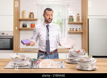 Un uomo triste con una pila di piatti sporchi in una cucina con una spugna Foto Stock