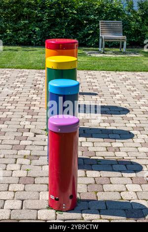 Una fila di vibranti strutture cilindriche in vari colori sorge su un sentiero lastricato accanto a una panchina del parco sotto cieli limpidi, creando un'atmosfera giocosa Foto Stock