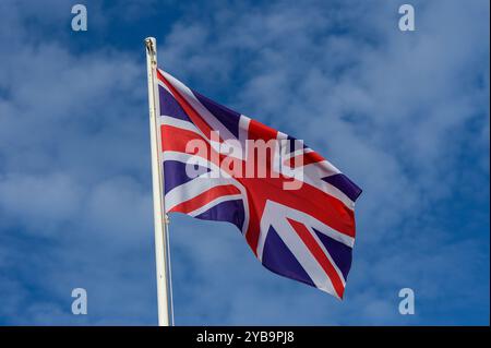 La colorata bandiera britannica sventola orgogliosamente in cima a un palo, con lo sfondo di un cielo blu vibrante punteggiato da soffici nuvole, catturando un momento di gioia Foto Stock