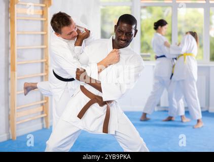 Classi di judo o jiu-jitsu - due uomini che praticano l'afferramento e il lancio di tappeti sportivi sotto la guida di un allenatore Foto Stock