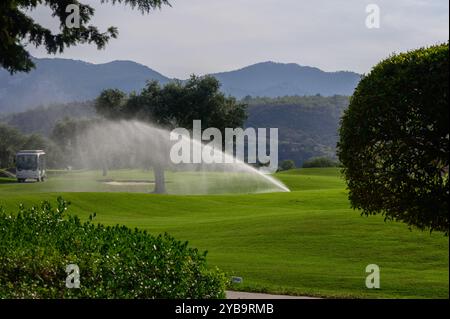 Un delicato getto di spruzzatori rivitalizzerà i verdi vibranti di un campo da golf in una mattinata di sole, con montagne maestose in lontananza che aumentano la t Foto Stock