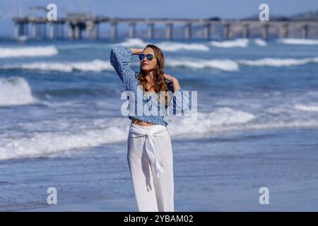 Una donna mozzafiato si crogiola nella beatitudine della spiaggia, vicino a un molo, sotto cieli azzurri, abbracciando la serenità di una giornata perfetta in riva al mare Foto Stock