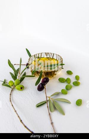 Olio d'oliva in una ciotola di cristallo, olive verdi e nere in rami e olive verdi sfuse, isolate in bianco. Foto Stock