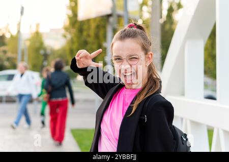 Una ragazza che dà un segno di pace mentre posa energicamente su una strada. Adolescente che scatta selfie mentre cammina lungo una strada della città la sera Foto Stock