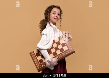 Una giovane ragazza che porta con sé una grande scacchiera di legno, guardando indietro con un sorriso. Foto Stock