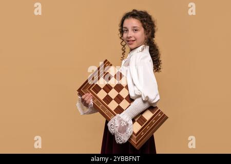 Ragazza giocatrice di scacchi che tiene una grande scacchiera sotto il braccio, sorridendo con fiducia. Foto Stock