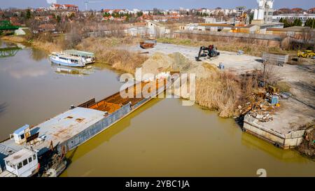 Vista dall'alto su escavatore, rimorchio e rimorchiatore mentre estraggono parte del letto di una vecchia nave merci, chiatta, lungo la costa del fiume, cassazione, nave da demolizione Foto Stock