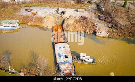 Vista dall'alto su escavatore, rimorchio e rimorchiatore mentre estraggono parte del letto di una vecchia nave merci, chiatta, lungo la costa del fiume, cassazione, nave da demolizione Foto Stock