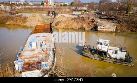 Vista dall'alto su escavatore, rimorchio e rimorchiatore mentre estraggono parte del letto di una vecchia nave merci, chiatta, lungo la costa del fiume, cassazione, nave da demolizione Foto Stock