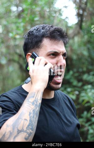 Un giovane frustrato urla nel suo cellulare durante un'intensa telefonata, in piedi in mezzo a una foresta pacifica Foto Stock