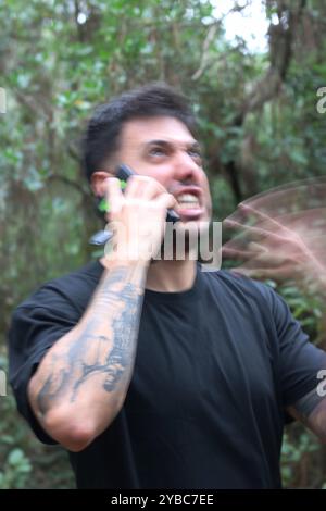 Un giovane frustrato urla nel suo cellulare durante un'intensa telefonata, in piedi in mezzo a una foresta pacifica Foto Stock