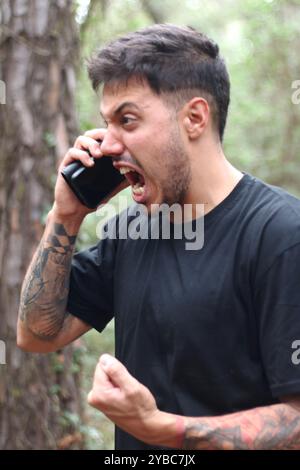 Un giovane frustrato urla nel suo cellulare durante un'intensa telefonata, in piedi in mezzo a una foresta pacifica Foto Stock