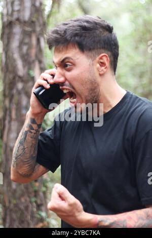 Un giovane frustrato urla nel suo cellulare durante un'intensa telefonata, in piedi in mezzo a una foresta pacifica Foto Stock