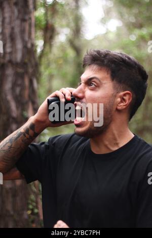 Un giovane frustrato urla nel suo cellulare durante un'intensa telefonata, in piedi in mezzo a una foresta pacifica Foto Stock